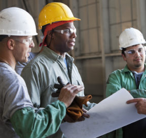 A group of four working men are wearing hard hats and work clothes as they discuss a plan on a white sheet of paper. Two of the men are wearing yellow hats, and the other two are wearing white hats. They are all wearing safety glasses. For page "Builder Development Program."